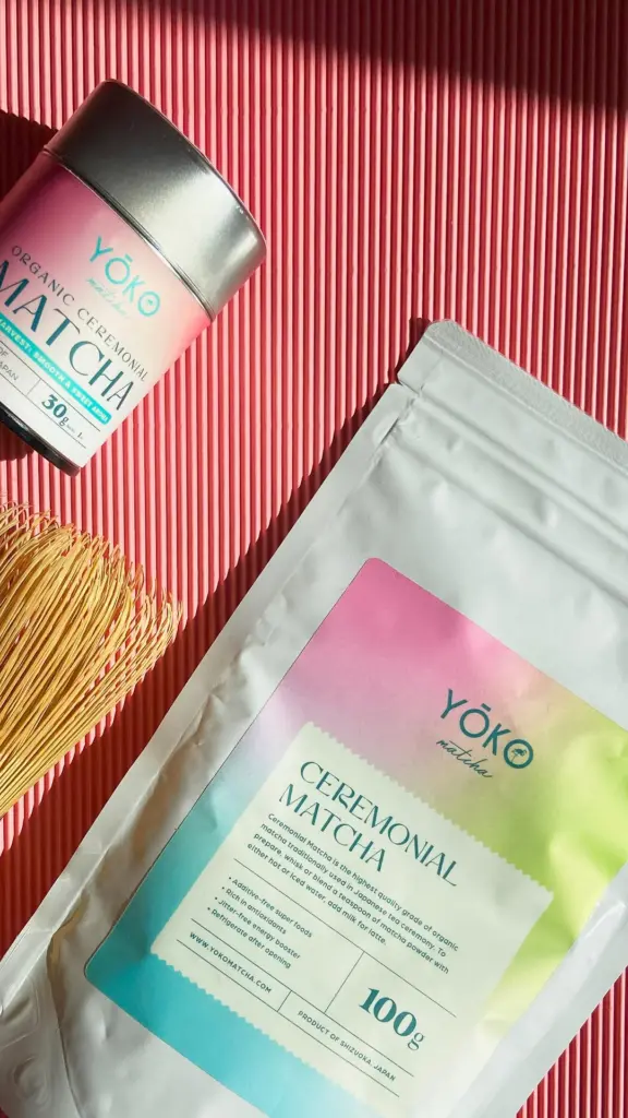 Yōko Matcha tin and pouch beside a bamboo whisk on a ribbed pink surface