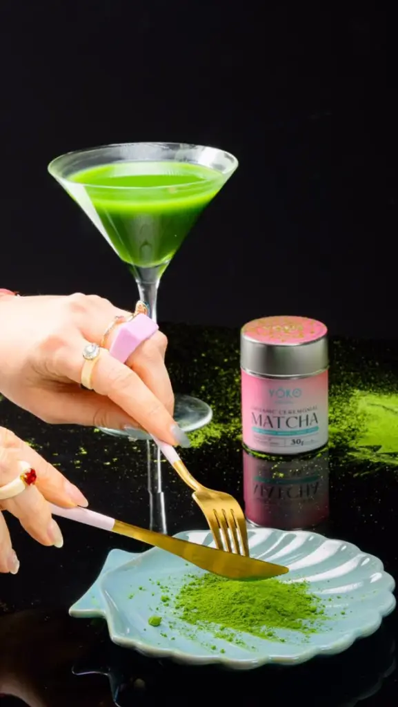Green drink in a stemmed glass beside a pink Yōko Matcha tin and matcha powder on a plate