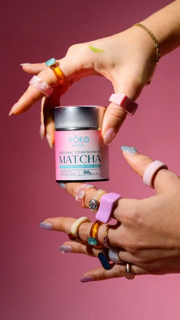Ringed hands holding a pink Yōko Matcha tin against a deep pink background