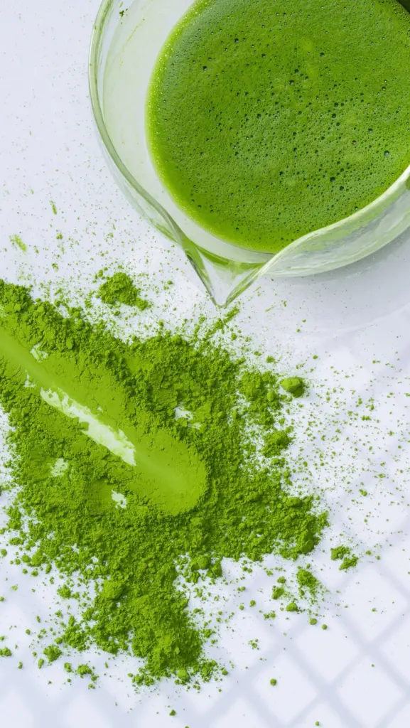 Foamy green matcha in a glass pitcher beside scattered bright green powder