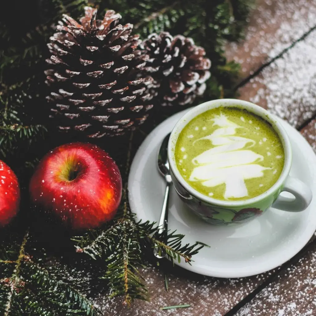 Matcha latte with tree-shaped foam art beside red apples, pinecones, and evergreen branches