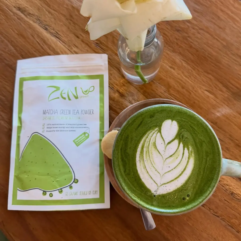 Matcha latte with leaf-shaped foam art beside a Zen Green Tea powder pouch