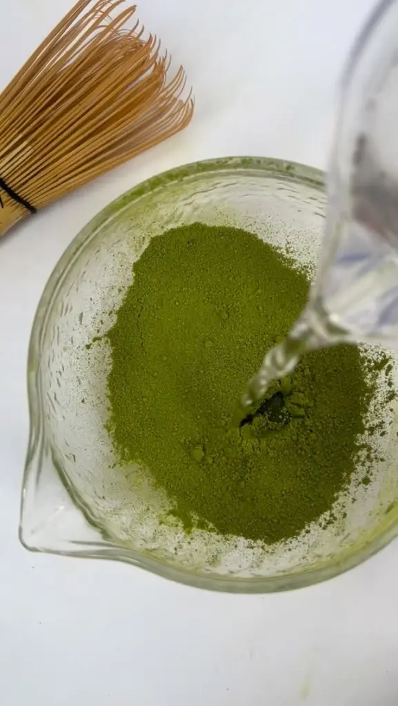 Water pouring into green matcha powder in a glass bowl beside a bamboo whisk